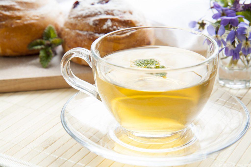 Cup of green tea with lemon balm and tasty rolls, blue spring flowers on light wooden background